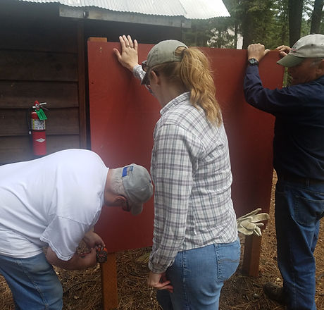 Two volunteers hold a wooden panel while one volunteer uses a power tool to secure it onto a post.