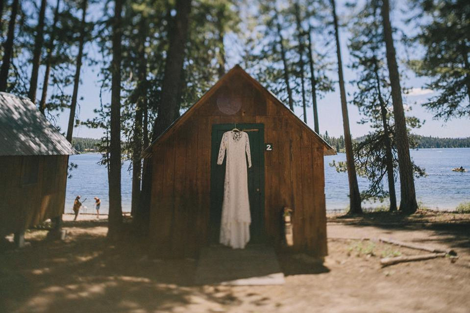 Wedding dress hangs on door in front of small cabin with lake and trees in background.