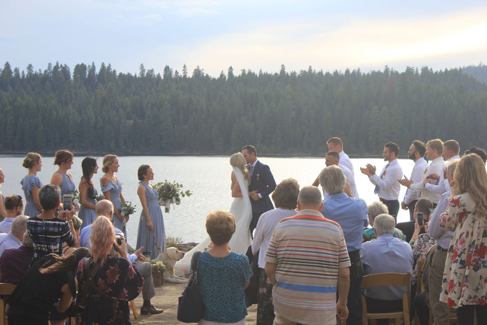 Wedding couple sharing first kiss while guests applaud at Paradise Point with lake in backgrounds.