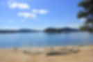 Empty campfire benches surround a sandy campfire area. In the background is a still lake with a view of Shellworth Island and blue skies.