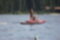 A camper with long hair and a red lifejacket paddles on a Stand Up Paddleboard away from the camera on a calm lake. A red kayak is in the background.
