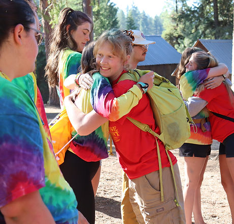 A camper in a red shirt hugs a staff member in a rainbow tie-dye shirt.