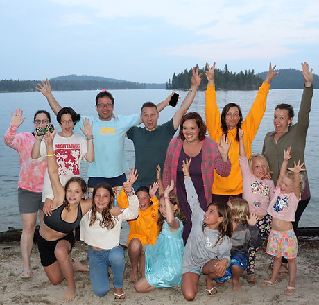 A family smiles with hands extended while standing in front of the Payette Lake on a sandy beach.