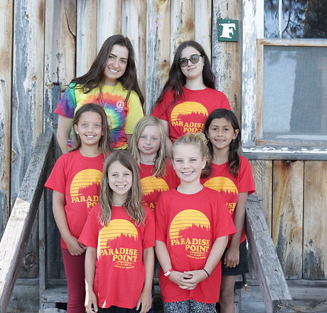 Youth campers in red shirts and their counselor wearing rainbow tie-dye smile in front of a rustic cabin.